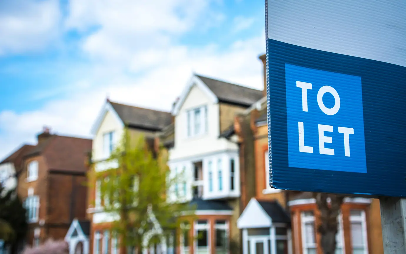 A blue to let sign, displayed in front of a row of read brick houses with white windows. 