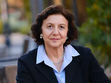 A woman in business attire is sitting at a table in a garden, her hands folded in front of her, looking into the camera.