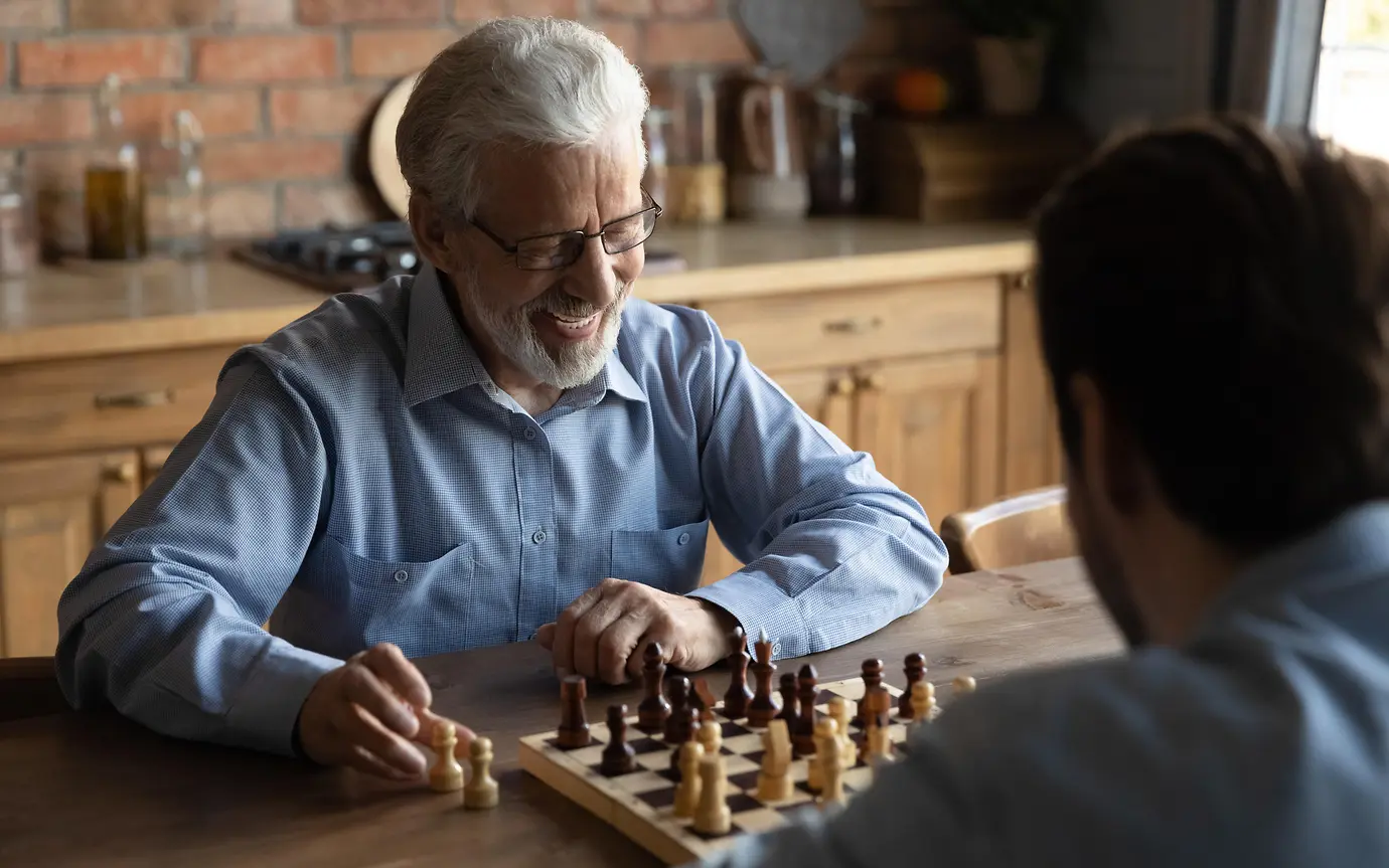 Smiling elderly Caucasian 60s father have fun play chess at home on weekend together with adult son.
